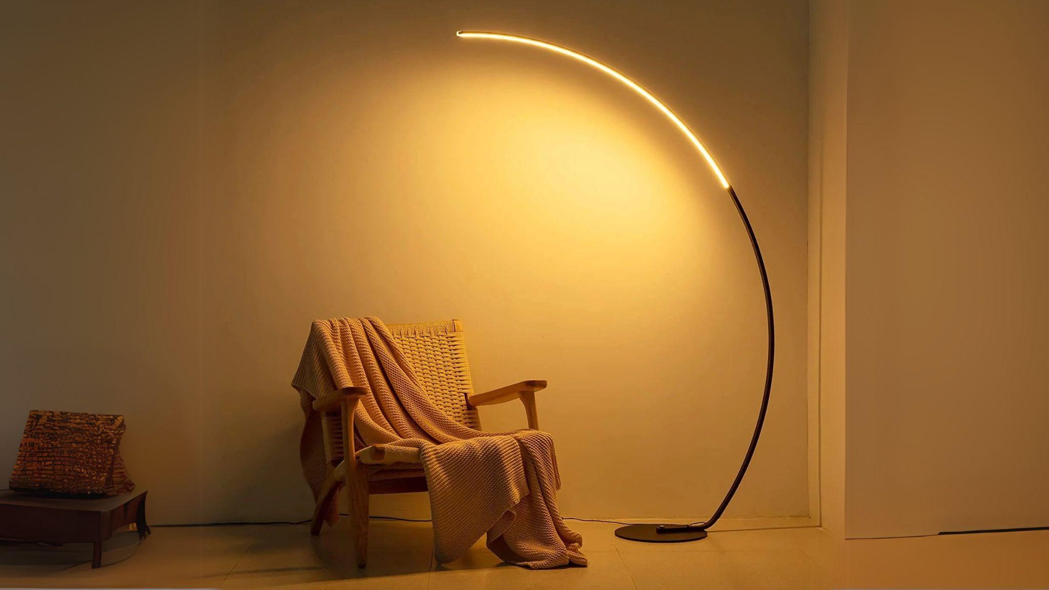 RGBW Modern Curve Floor Lamp casting warm LED light over cozy chair with a blanket in a minimalist room.