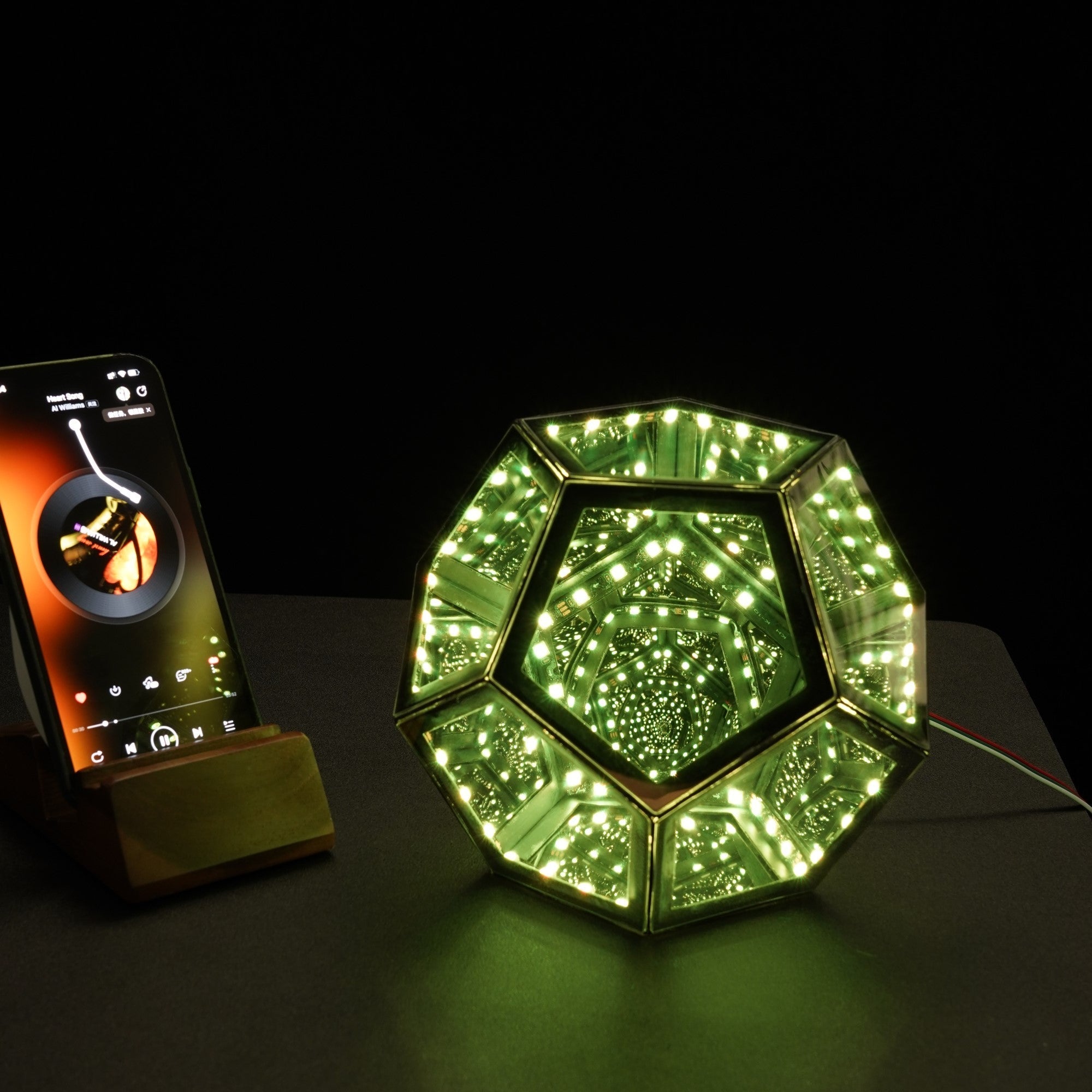Geometric Infinity Lamp (RGB) glowing green with infinite light tunnel effect on a dark table beside a smartphone.