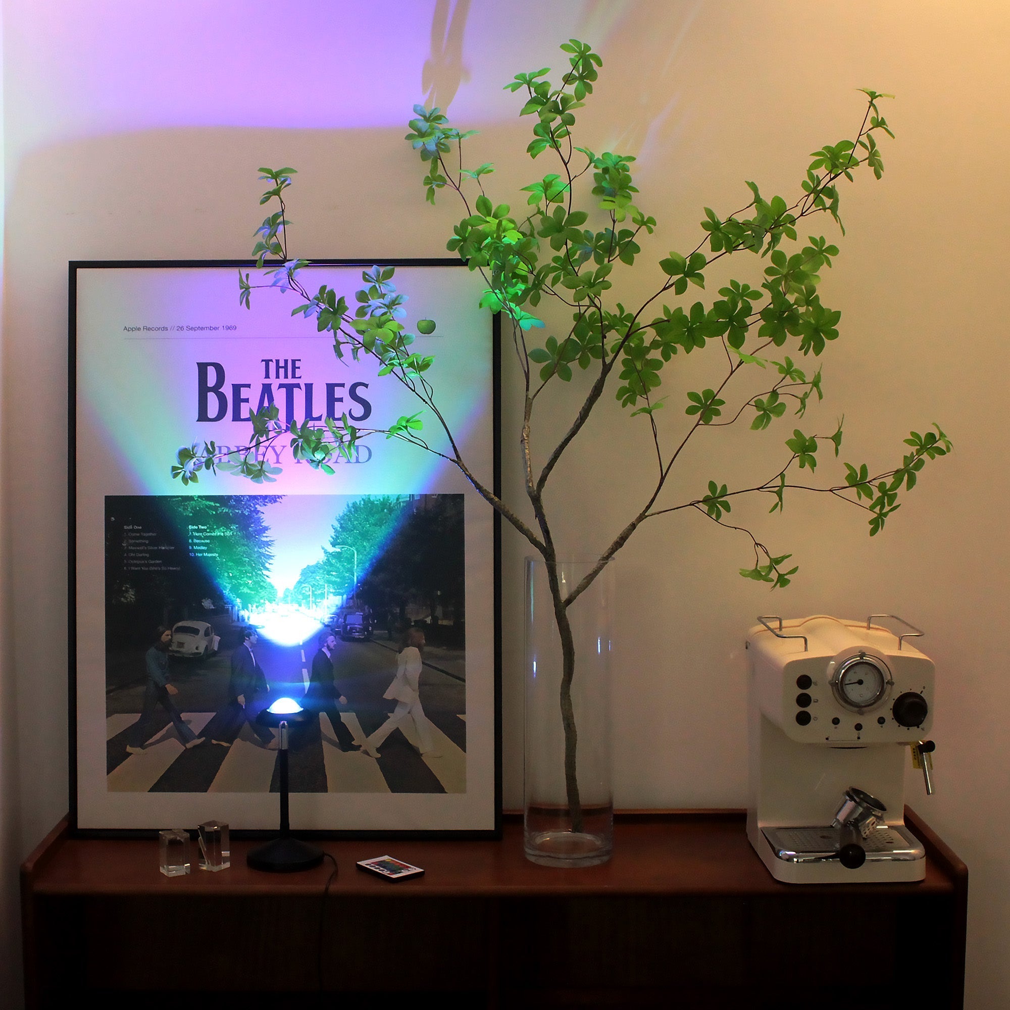RGB Sunset Projector Lamp casting colorful light on a Beatles poster, creating vibrant ambiance on a wooden shelf.