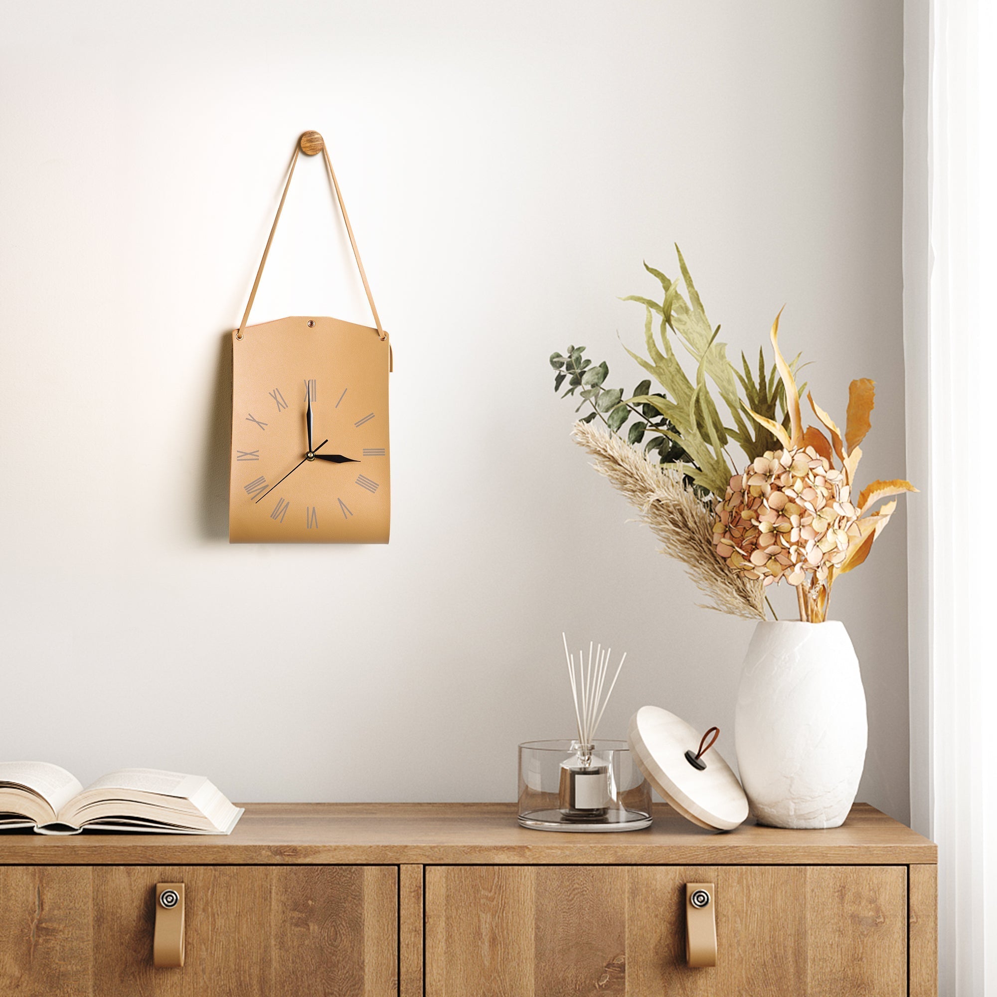 Roman Numerals Leather Wall Clock hanging on wall above wooden cabinet with decorative vase and open book.